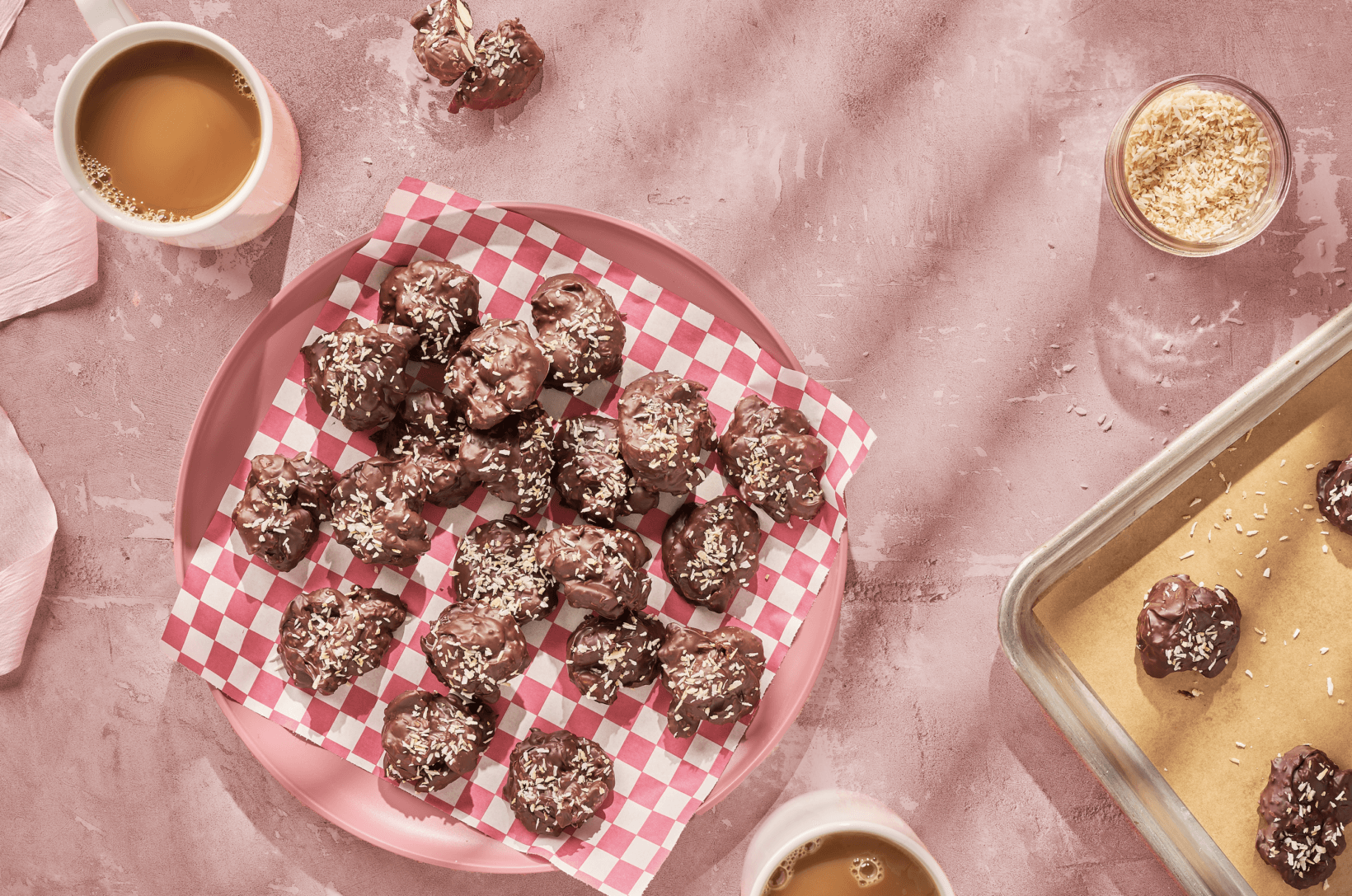 Chocolate almond clusters on checked paper on a pink plate, with coffee and a tray in the background on a pink surface.