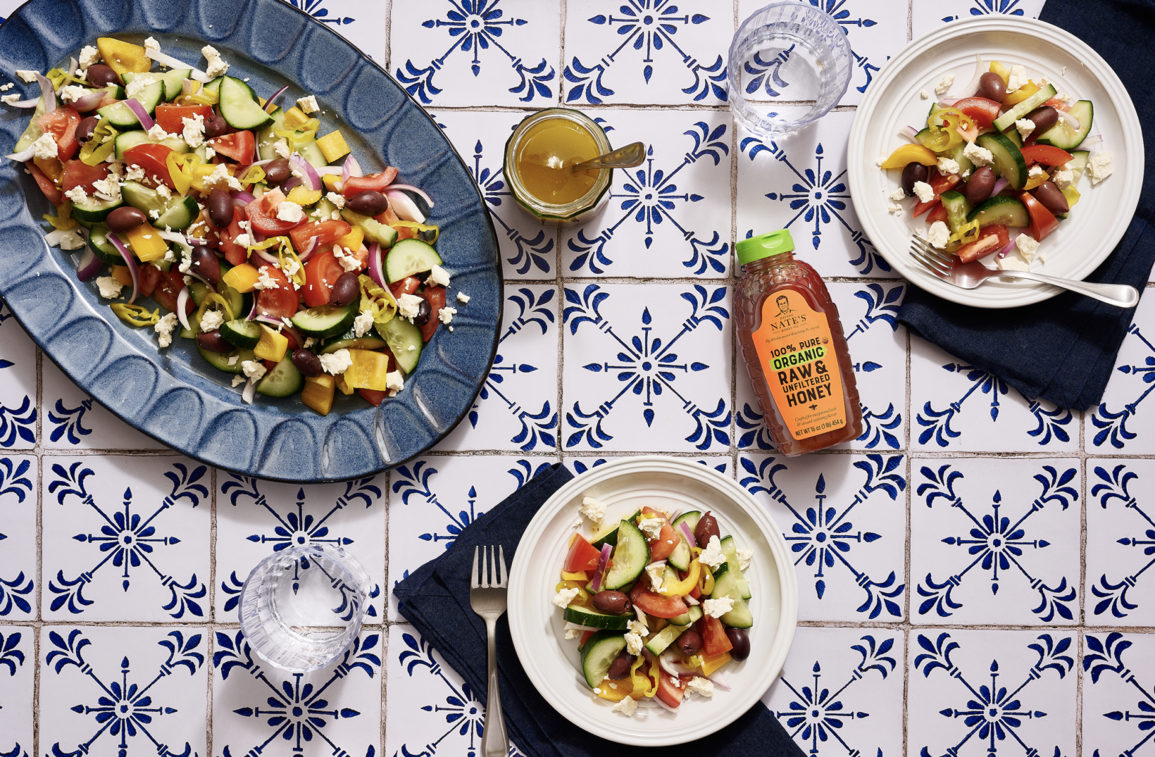 Greek salads, honey salad dressing and a bottle of Nate’s organic honey on a blue and white tiled table.