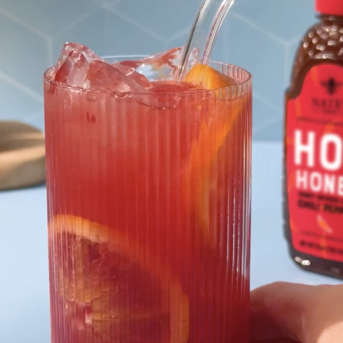 Glass of blood orange cocktail with ice and orange slices and Nate's hot honey bottle in the background.