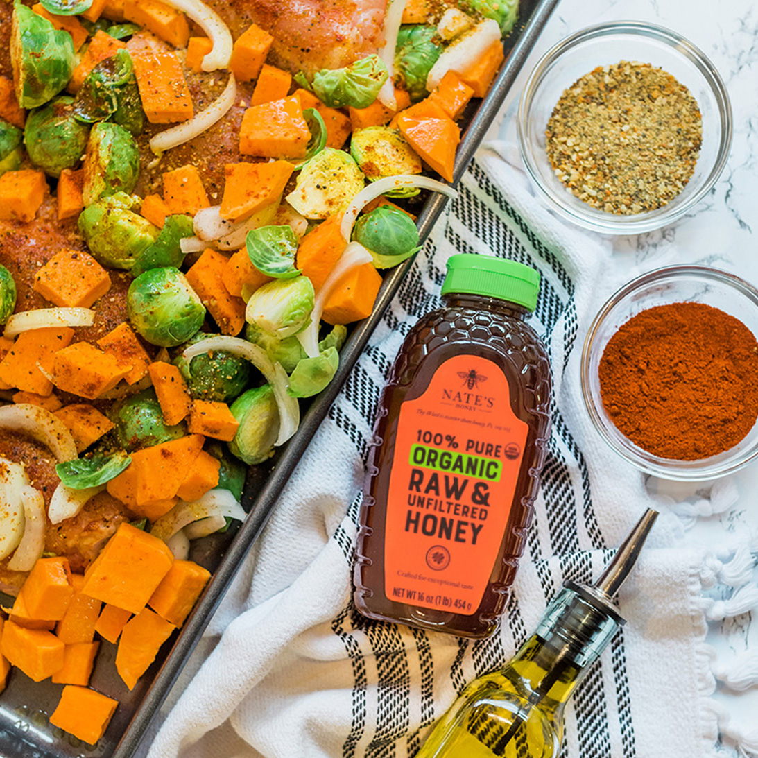 Nate’s organic honey bottle with roasted veggies, spices and olive oil on a striped cloth.