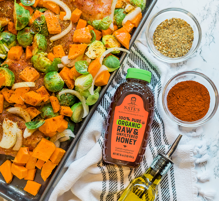 Nate’s organic honey bottle with roasted veggies, spices and olive oil on a striped cloth.