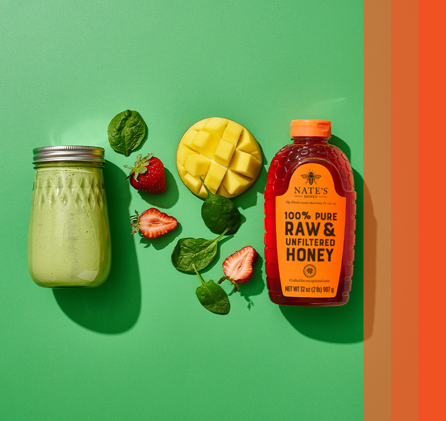 Mason jar filled with green liquid next to spinach leaves, strawberries, mango and Nate's organic honey bottle.
