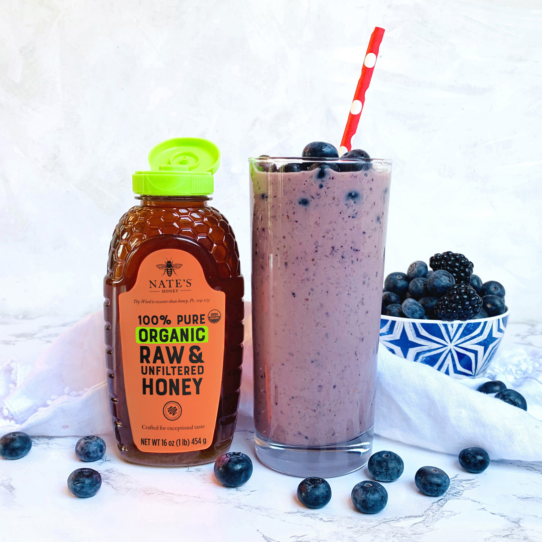 Blueberry smoothie with straw, Nate's Honey bottle, and bowl of berries on a marbled background.