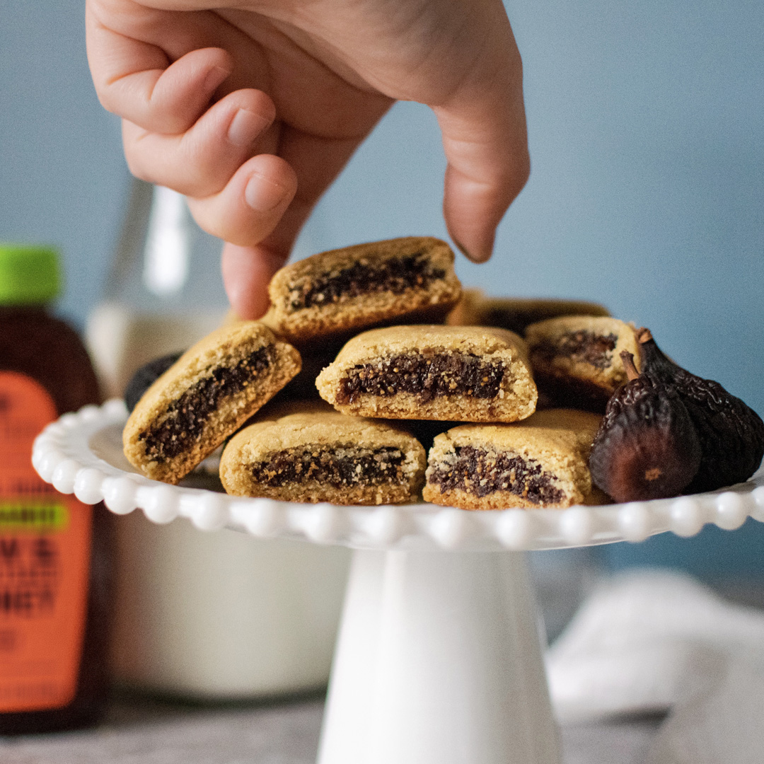 A hand taking a fig-filled cookie from a stack on a pedestal dish, with a bottle of Nate’s Honey in the background.