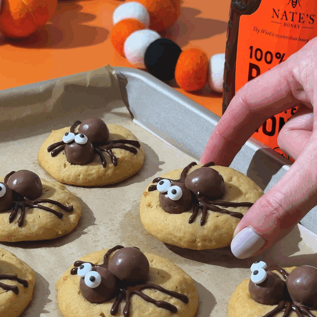 Spider cookies with chocolate bodies and candy eyes on a baking sheet, with a hand picking one up.