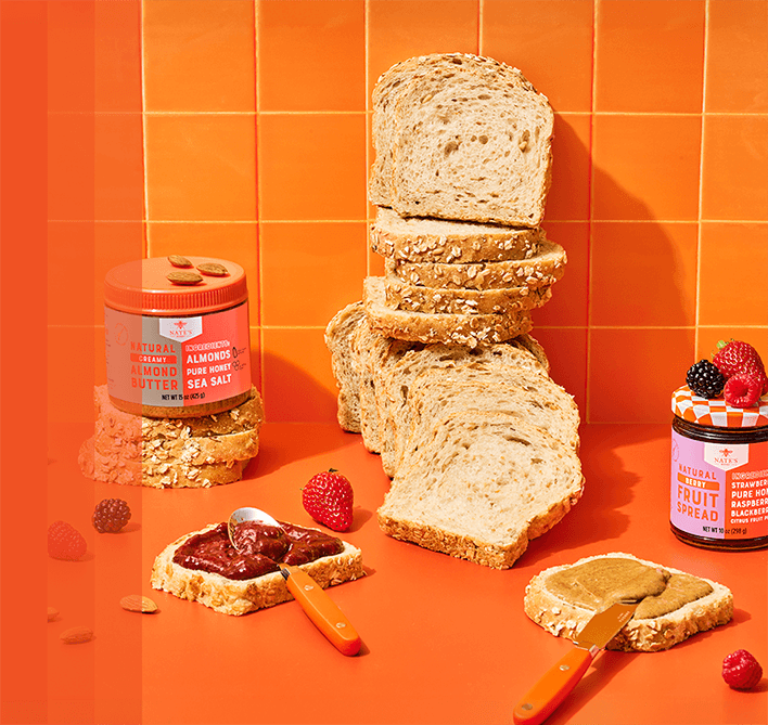 Nate’s Honey almond butter and berry fruit spread displayed with sliced bread, toast topped with fruit spread and almond butter, fresh berries and almonds on a bright orange background.