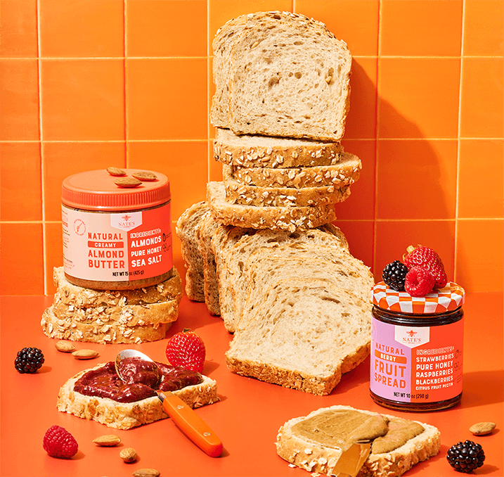 Nate’s Honey almond butter and berry fruit spread displayed with sliced bread, toast topped with fruit spread and almond butter, fresh berries and almonds on a bright orange background.