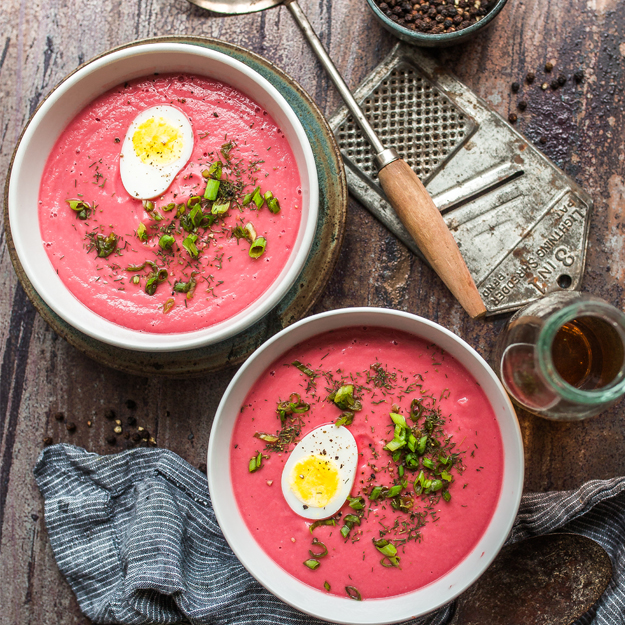Two bowls of beet and cauliflower soup, garnished with eggs and chives, rustic kitchen setting.
