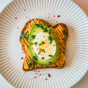 Hummus, avocado and egg toast with red pepper flakes and parsley on a white striped plate.