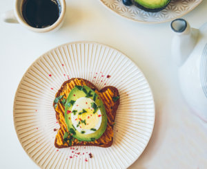 A poached egg atop avocado toast on a speckled plate, with half an avocado and tea set nearby.