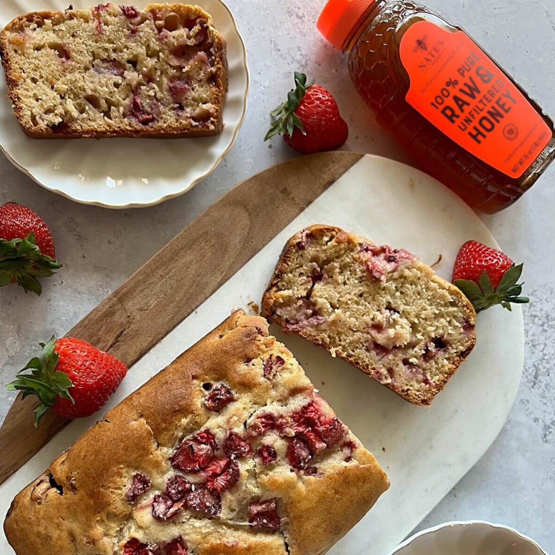 Sliced strawberry bread cake with a bottle of Nate’s honey on a marble countertop.