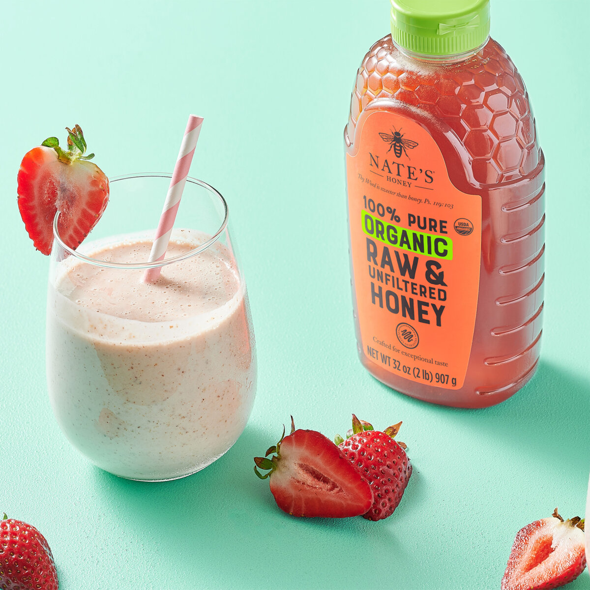 Nate's Organic Honey bottle next to glass of strawberry banana smoothie and slices of strawberries on teal background.