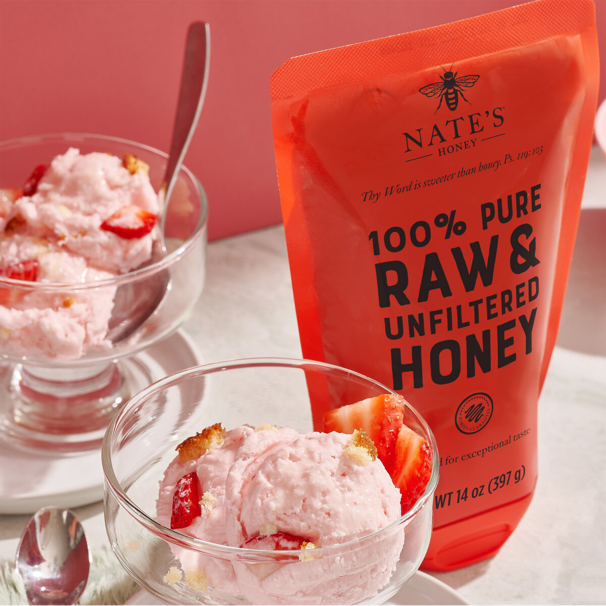 Strawberry ice cream in clear glass bowls drizzled with honey, next to a pouch of Nate’s Honey.