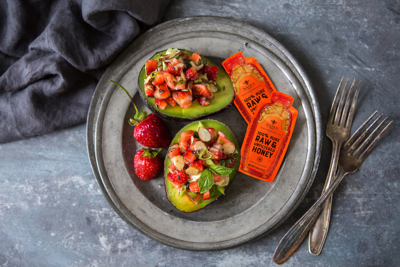 Avocado halves filled with strawberries and almonds on a metal plate with Nate's Honey Minis, beside a dark napkin.