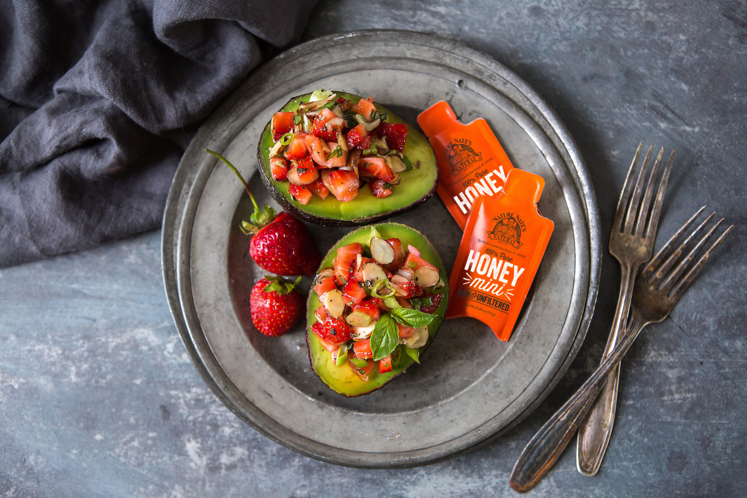 Avocado halves filled with strawberries and almonds on a metal plate with Nate's honey minis, beside a dark napkin.