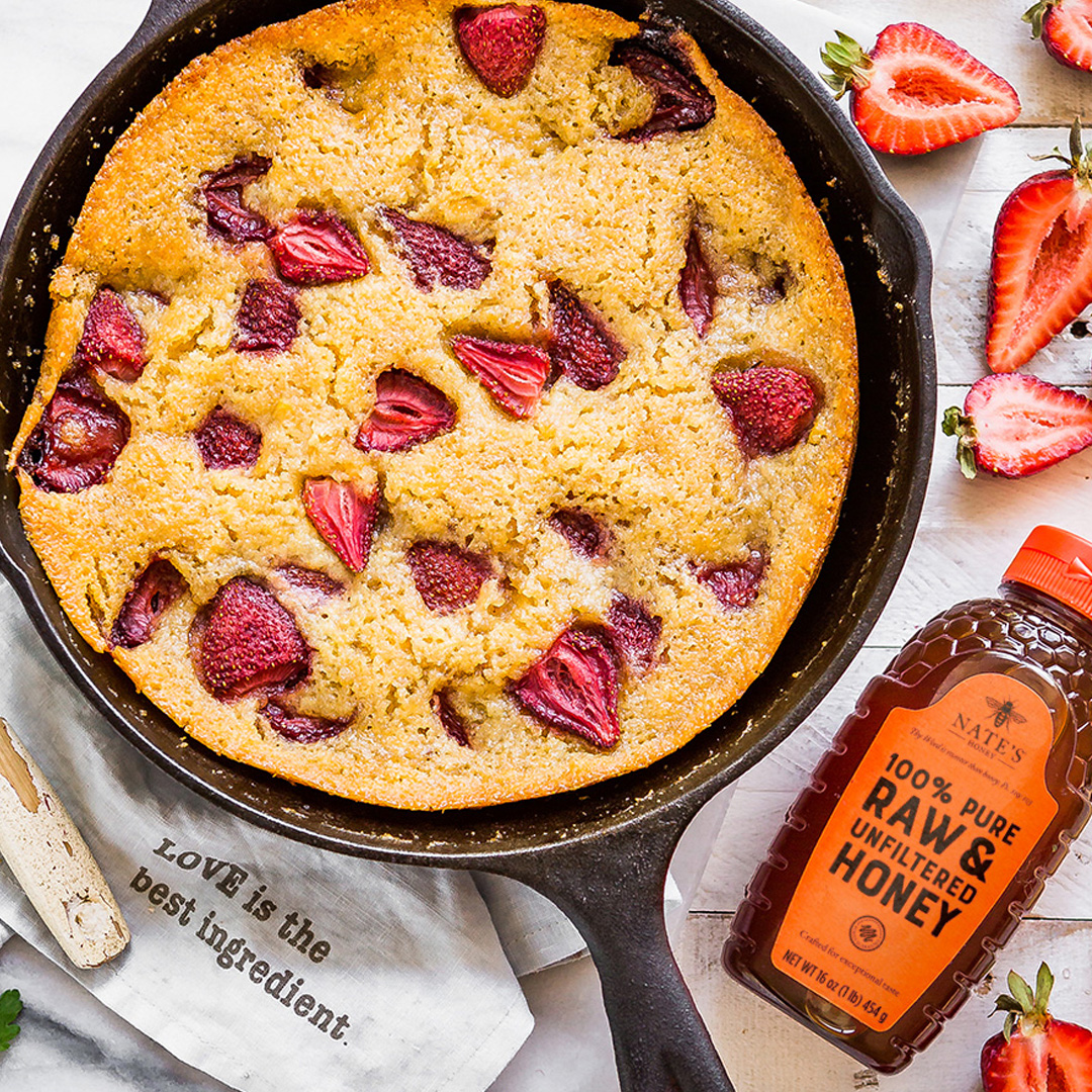 Strawberry corn cake in a skillet with a bottle of Nate's Honey and kitchen utensils, with strawberries and herbs.