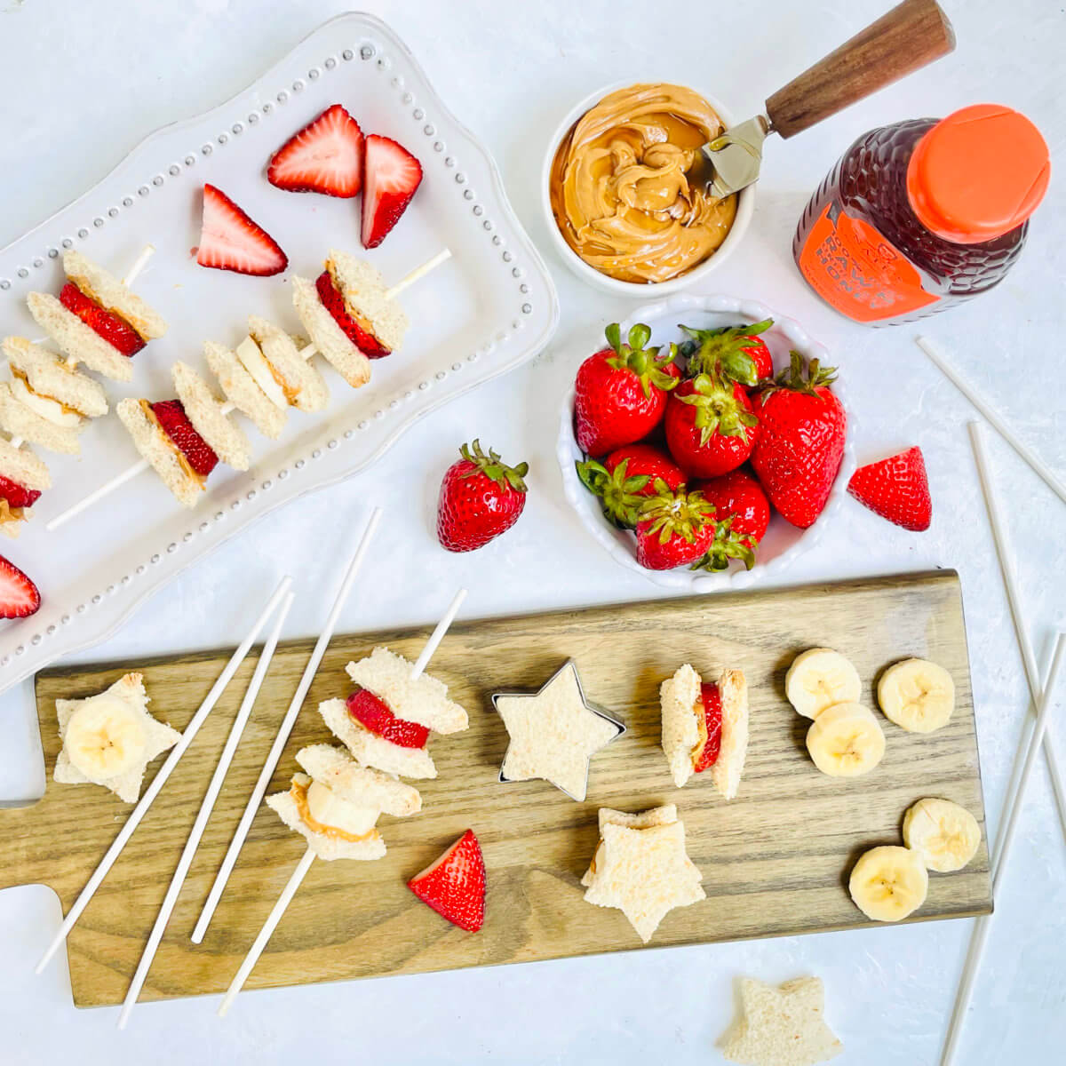 A snack setup with star-shaped sandwich skewers, peanut butter, Nate’s honey bottle and fresh strawberries on a board.
