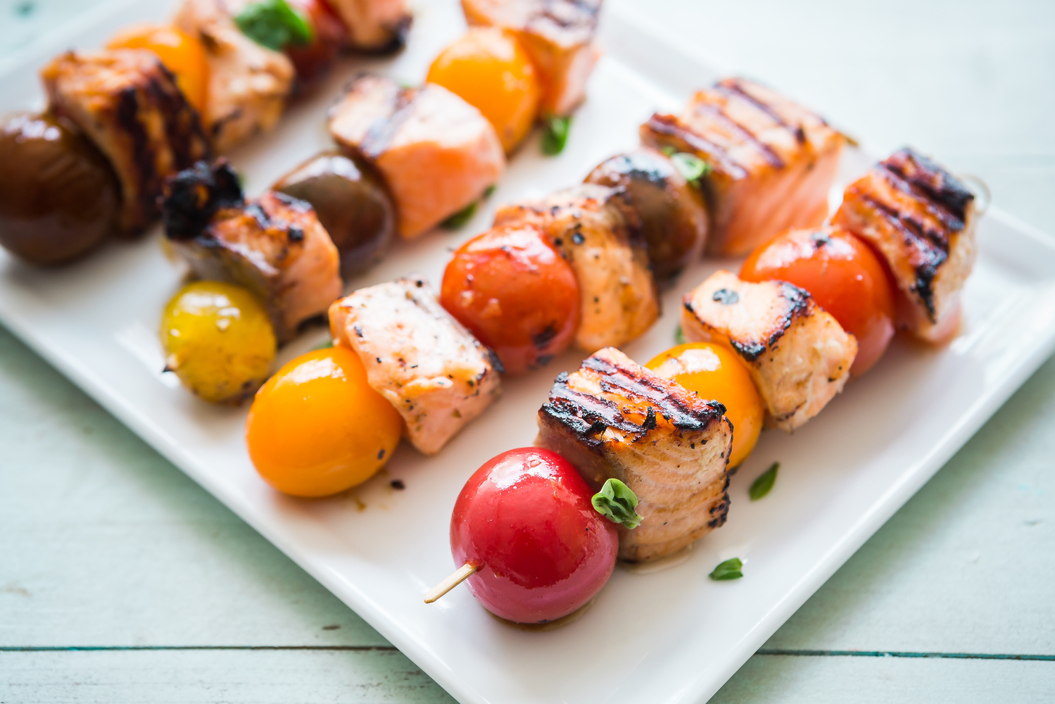Grilled skewers with salmon and colorful tomatoes on a white plate, wooden background.