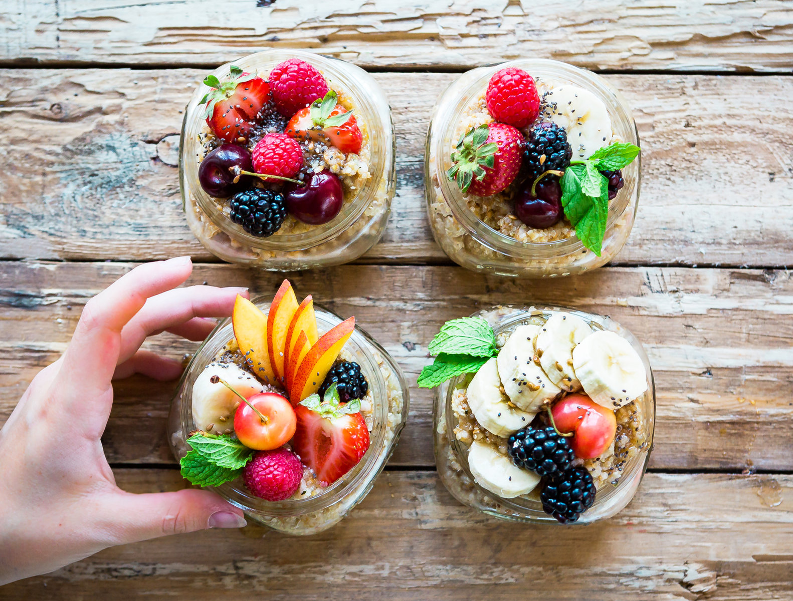 A hand holding one of four bowls of oatmeal topped with a variety of fresh fruits on a rustic wooden table.