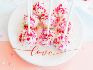 White chocolate-dipped cake pops with pink sprinkles on a plate with love script.

