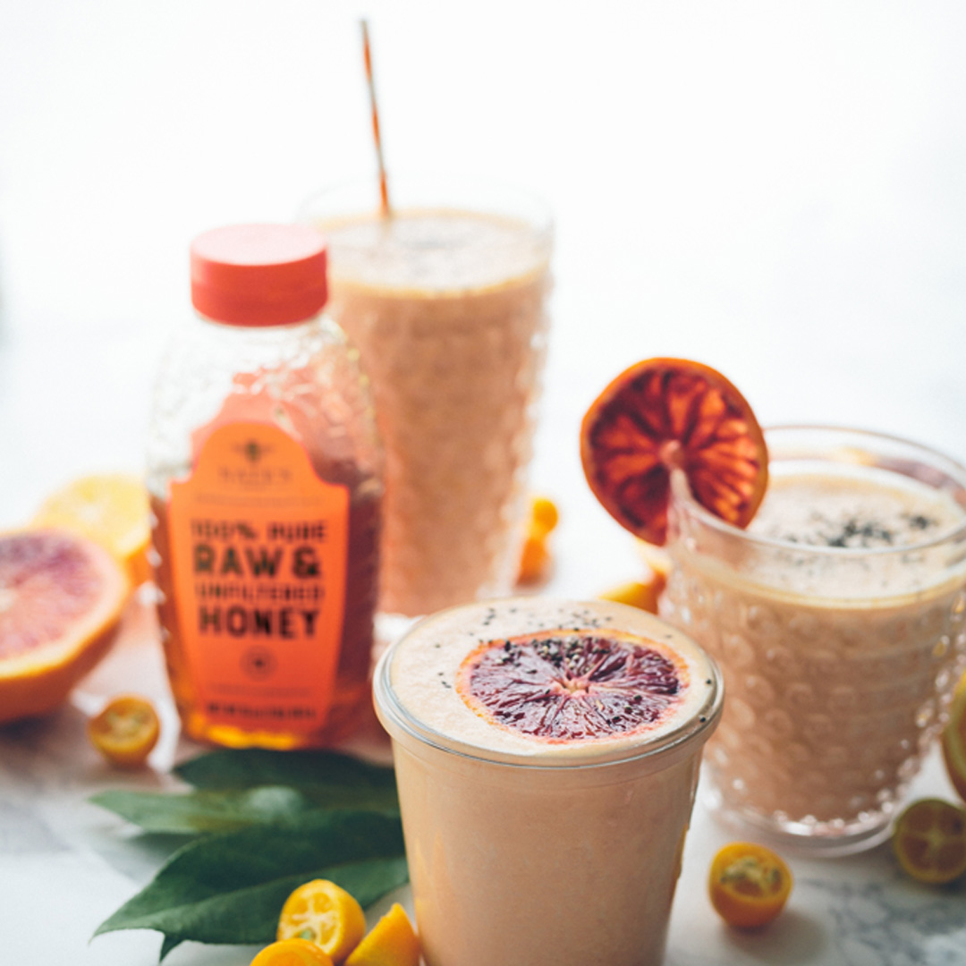 Sweet potato and orange smoothies with a bottle of Nate's Honey and fresh fruits on a marble surface.