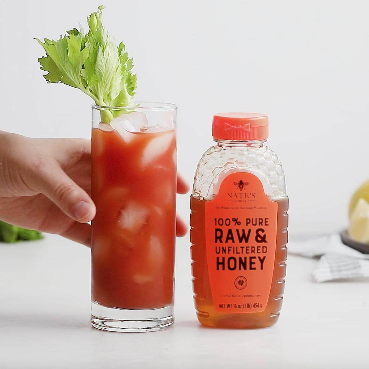 Hand holding a Bloody Mary cocktail with celery, next to a bottle of Nate’s Honey on a white background.