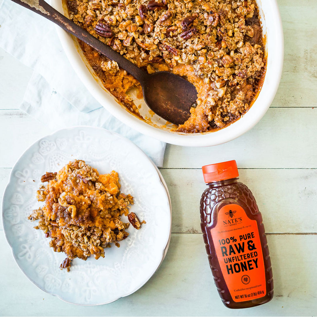 Baked sweet potato casserole in a baking dish and served on a plate and a bottle of Nate's honey on a wooden surface.