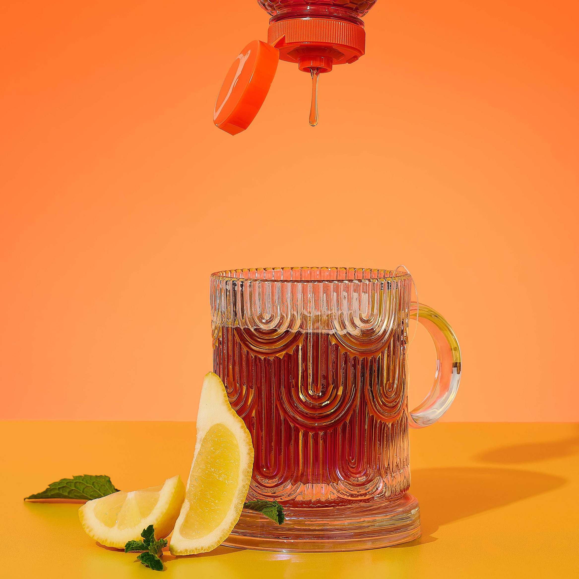 Honey dripping from squeeze bottle into a glass of tea with lemon wedges and mint on an orange background.