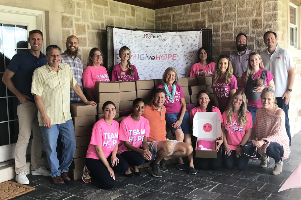 A volunteer group in pink shirts stands with a #GiveHOPE sign, surrounded by donation boxes.
