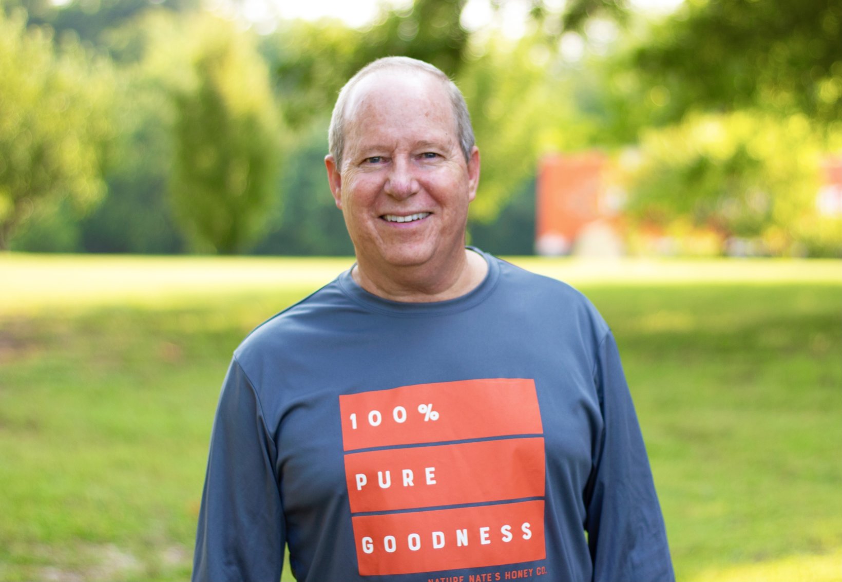 A smiling man in a park wearing a shirt with 100% PURE GOODNESS Nature Nate's Honey Co.