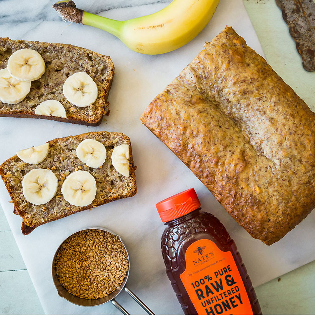 Banana bread with slices cut and topped with banana slices with Nate's Honey bottle on a light marble surface.