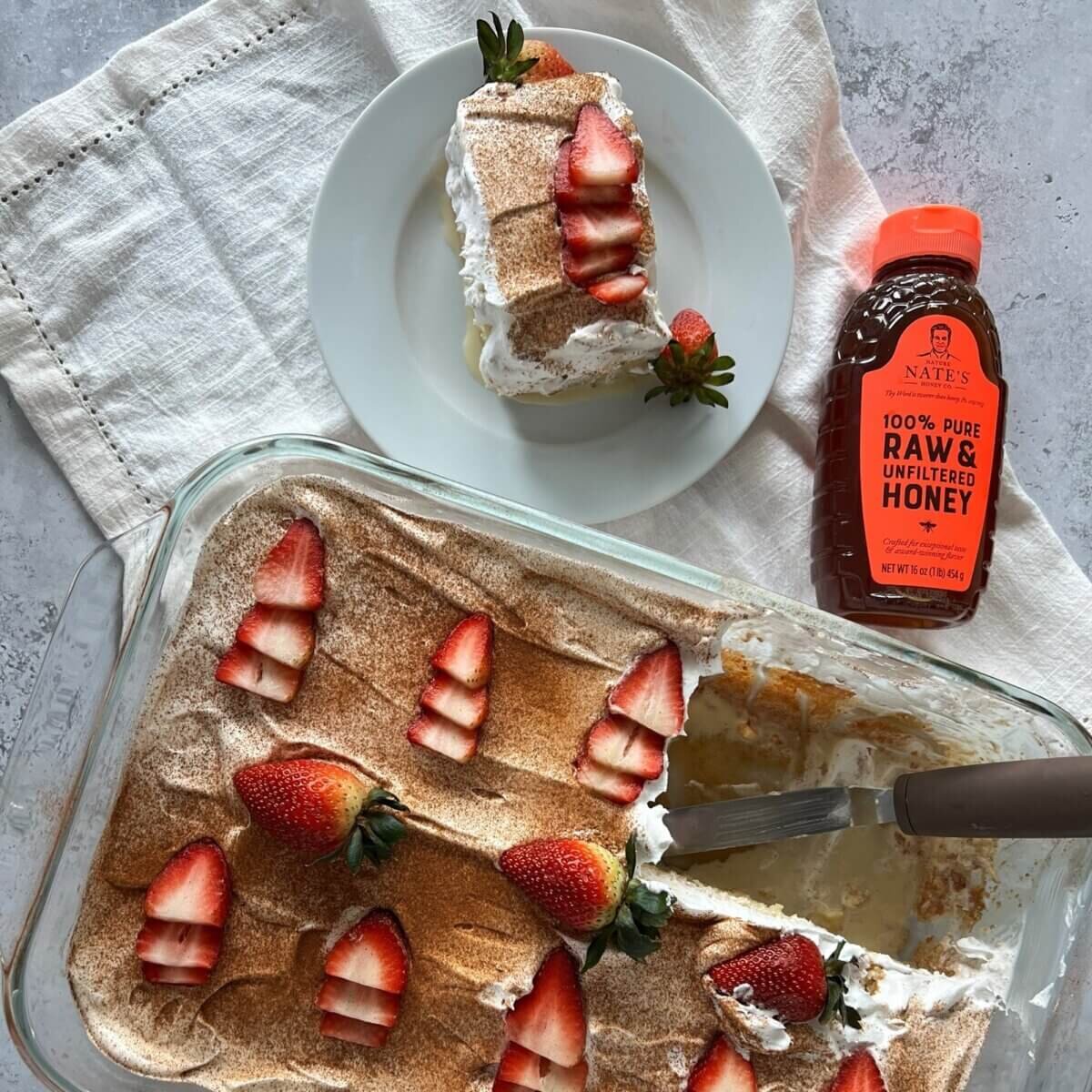 Tres leches cake with white frosting and garnished with strawberries and cocoa powder, next to a bottle of Nate's honey.