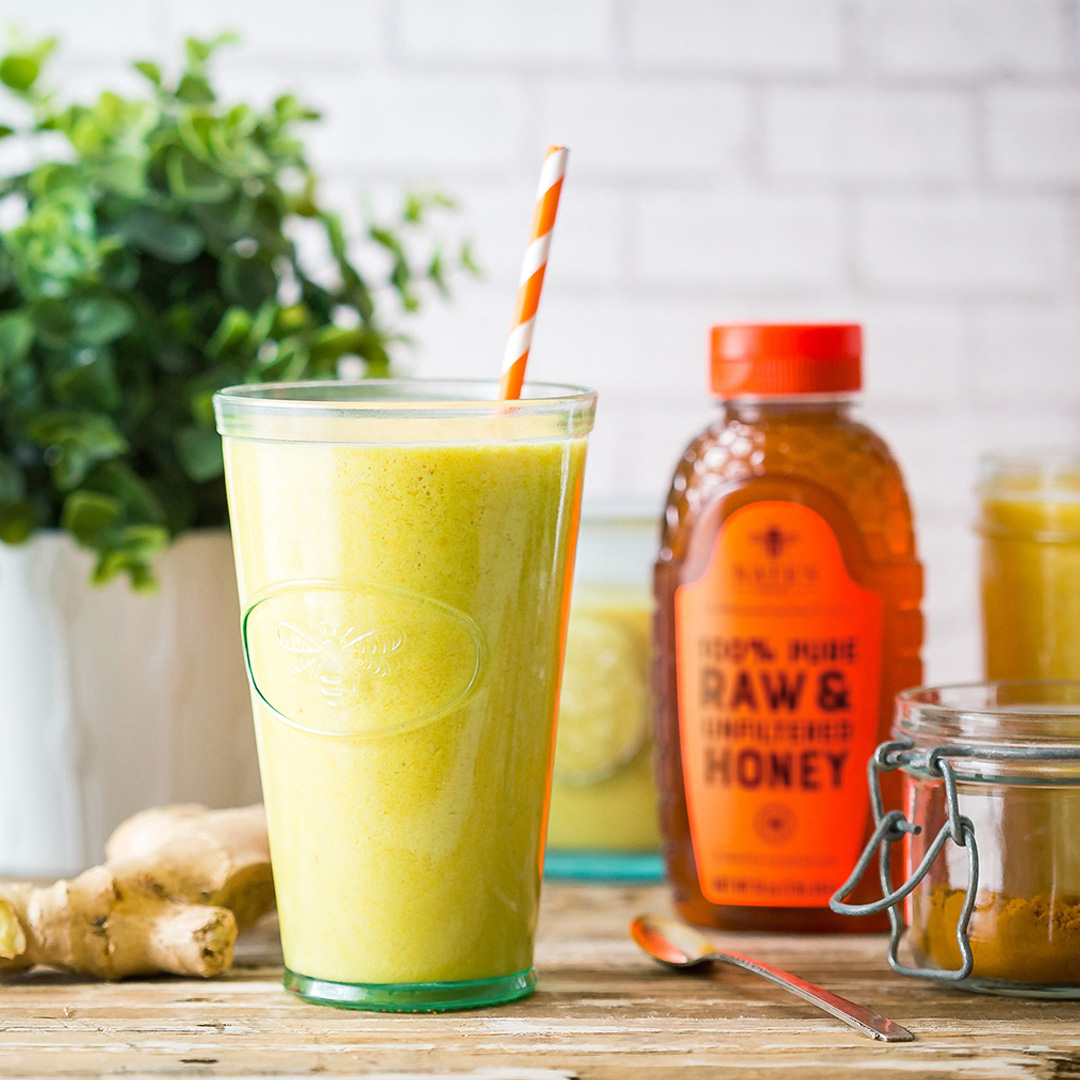 Green turmeric and ginger smoothie with a straw next to a bottle of Nate's Honey on a wooden table.