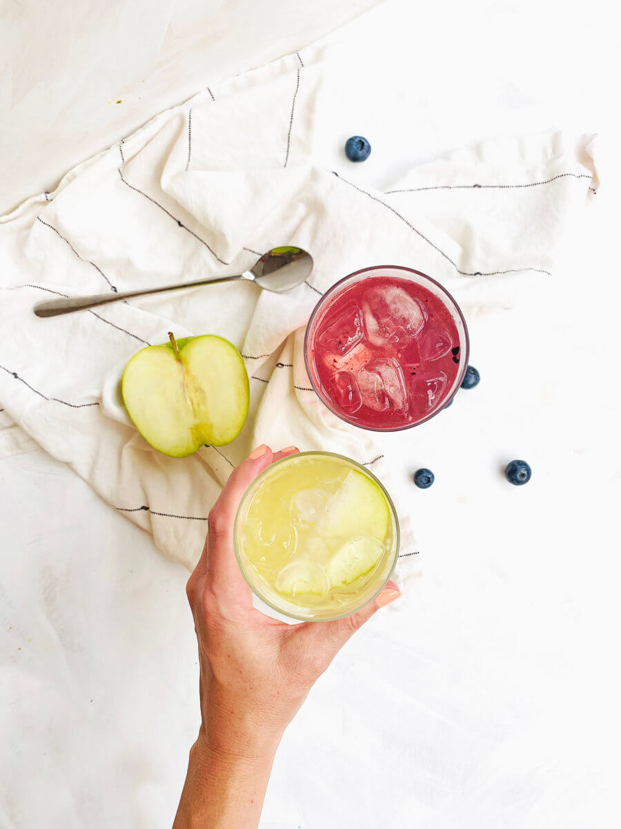 A hand holding a glass of a honey-lime drink, a sliced apple, a glass of a pink drink and blueberries on a white cloth.