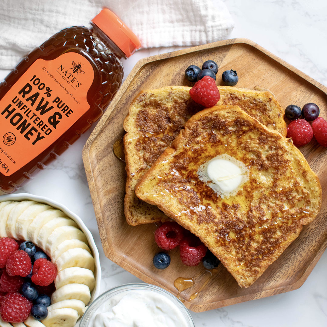 French toast with berries and banana on a plate, Nate’s Honey bottle with an orange cap, on a marble background.