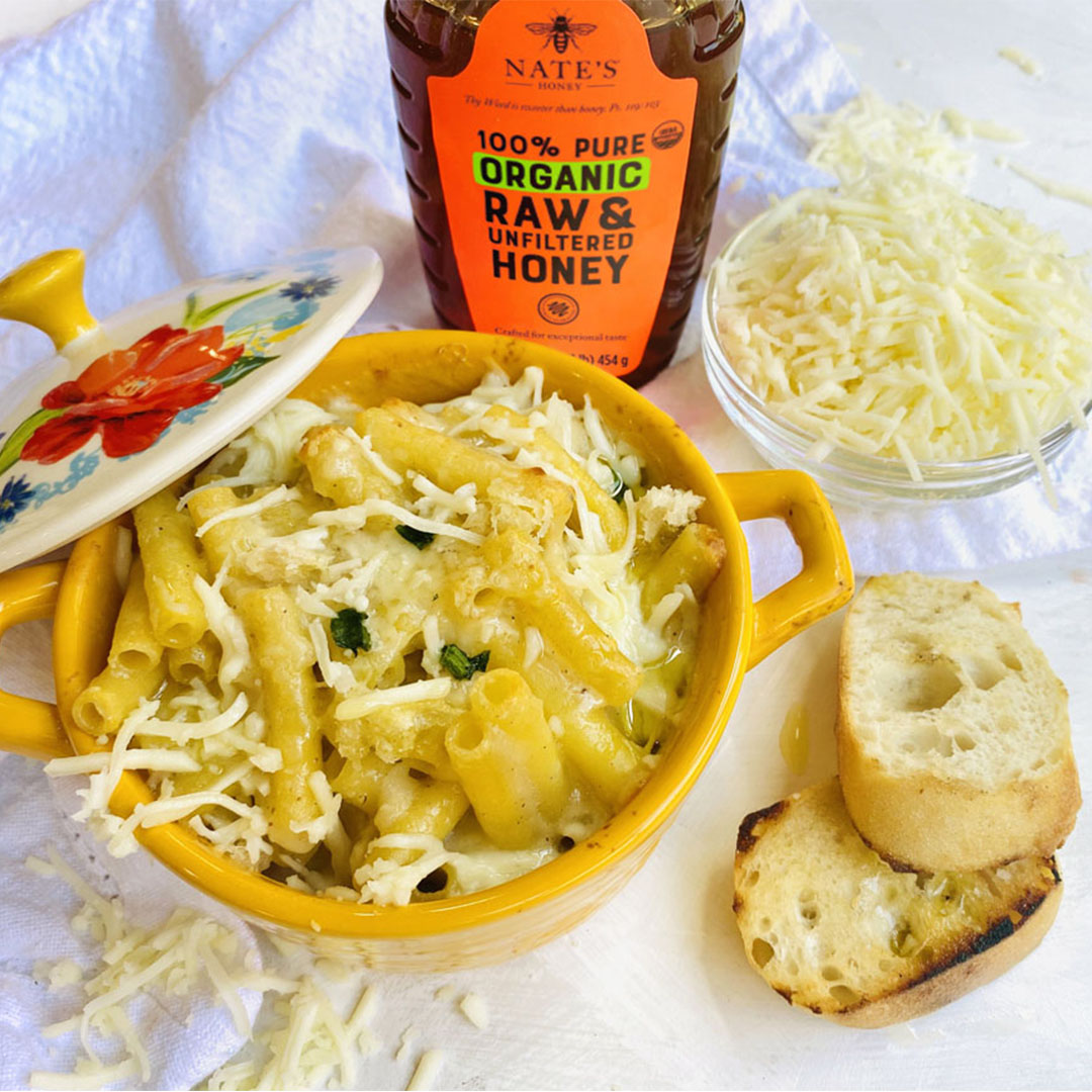 Cheesy pasta in a yellow dish with a bottle of Nate’s Honey, shredded cheese and garlic bread on crinkled white cloth.