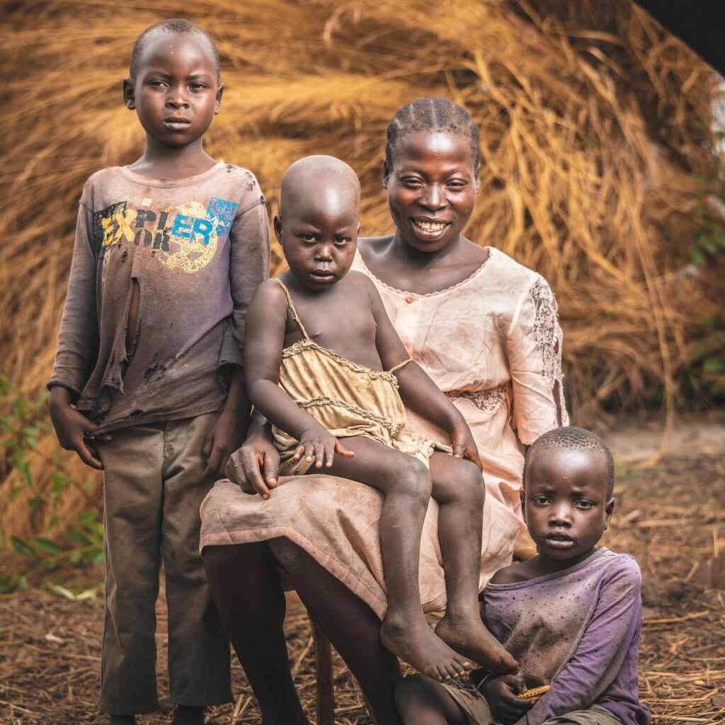 A woman and three children posing with warm smiles, in front of a thatched structure.