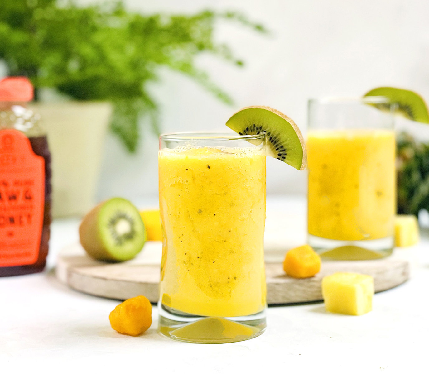 Glasses of fresh mango smoothie with kiwi slices, accompanied by a Nate's honey bottle and fruits on a light background.