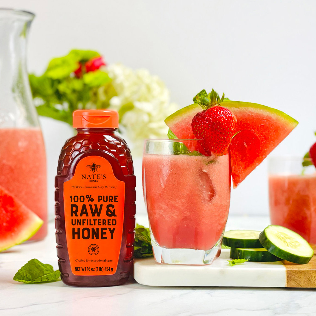 Watermelon slushies with lime, a bottle of Nate’s honey, and watermelon and cucumber slices on a marble counter.