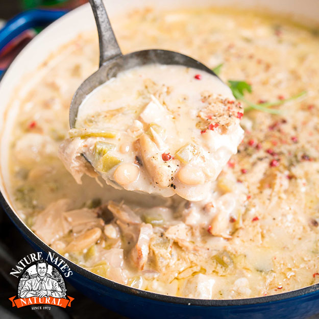 A ladle of creamy chicken soup, garnished with herbs, in a pot beside the Nature Nate's honey logo.