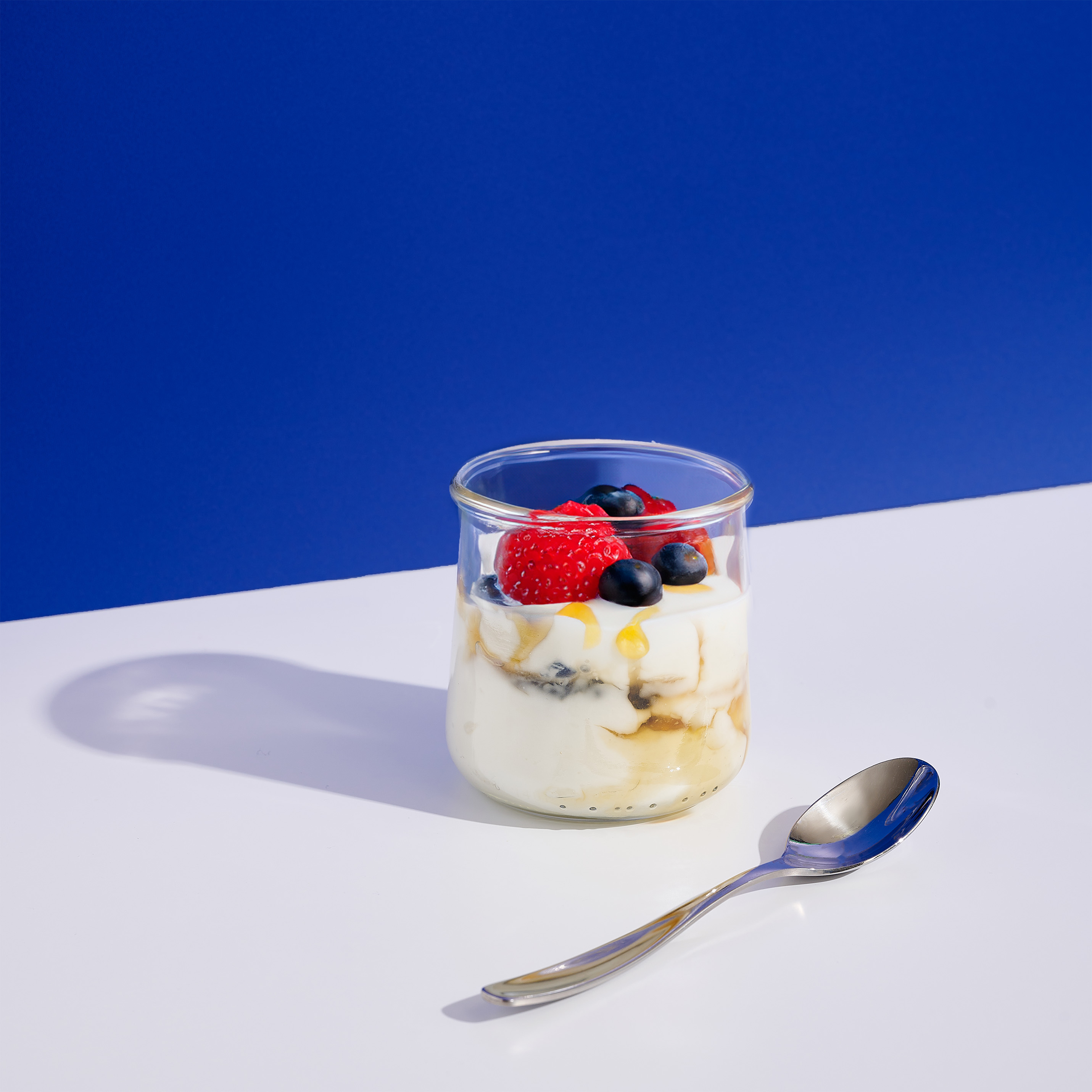 Clear jar of yogurt topped with honey and berries on a blue background.