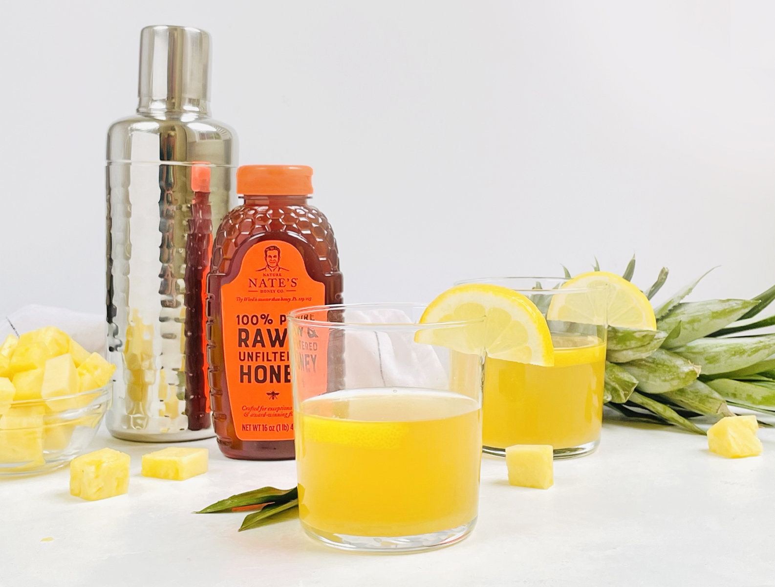 Pineapple cocktails with lemon garnish, Nate’s honey bottle, cocktail shaker and pineapple pieces.