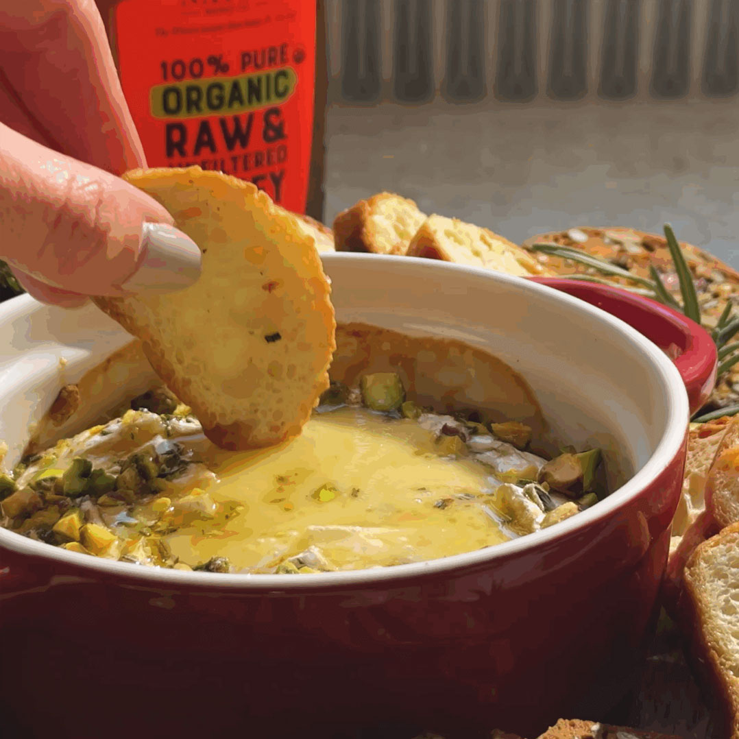 Hand dipping bread into bowl of baked brie with rosemary and pistachios with more bread and bottle of Nate's Organic Honey in background