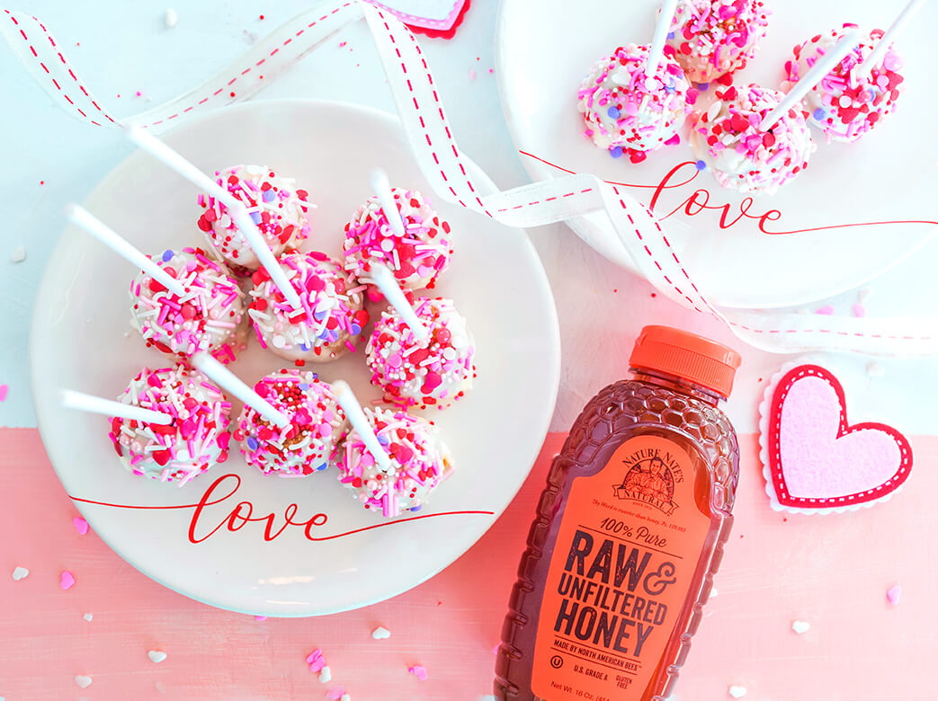white plates with cake pops with pink sprinkles next to bottle of Nate's honey and heart on pink and white table