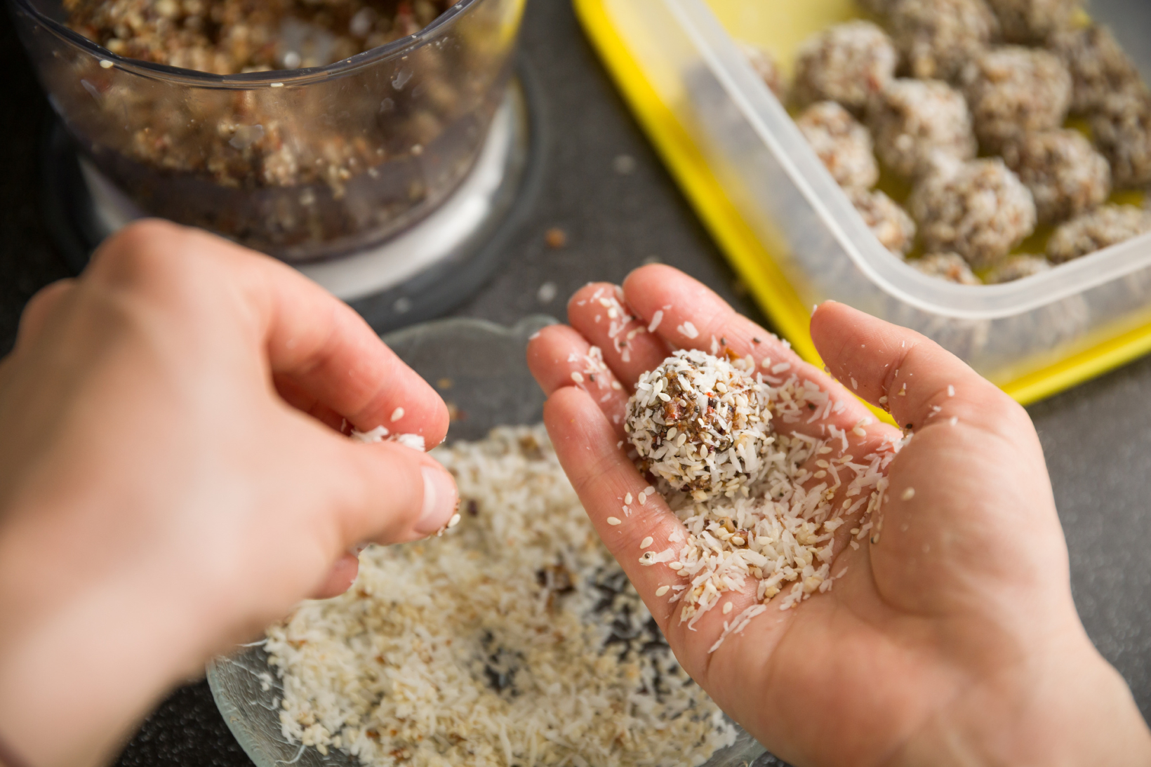 Hands making energy bites with coconut flakes, blurred background of ingredients and tray.