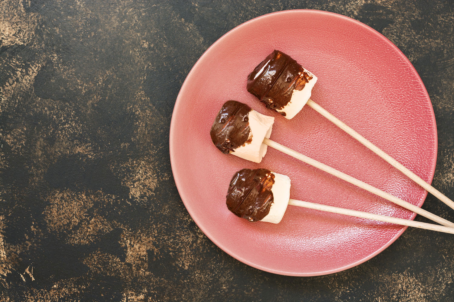 Marshmallows dipped in chocolate on sticks, presented on a pink plate with a dark textured backdrop.