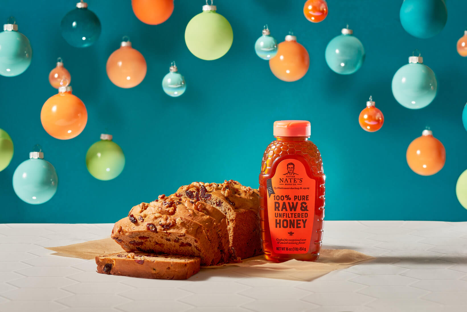 Cranberry nut bread beside a Nate’s honey bottle, against a backdrop with colorful hanging ornaments.