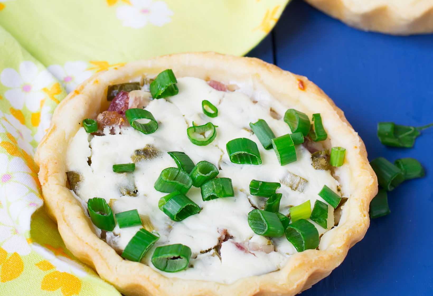 A savory tart with cheese and bacon filling and chopped green onions on top, on a blue surface with a floral napkin.