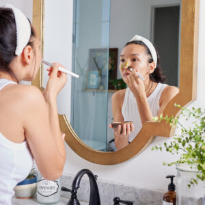 A woman at a bathroom sink with a bottle of Nate's honey, looking in the mirror. Person applying a natural face mask at a bathroom mirror as part of a skincare routine.
