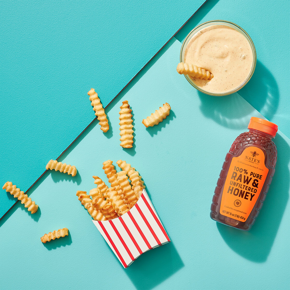 Fries spilling from a striped box with dipping sauce and a bottle of Nate’s honey on a teal background.